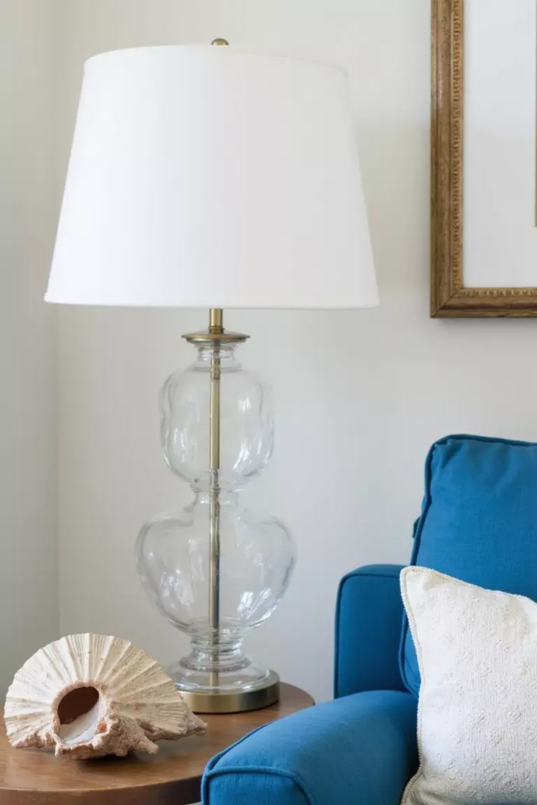 A professional photo, similar to a photo in a home design magazine, of a coastal living room corner with a clear glass lamp base, white shade, blue sofa, and seashell decor nearby.