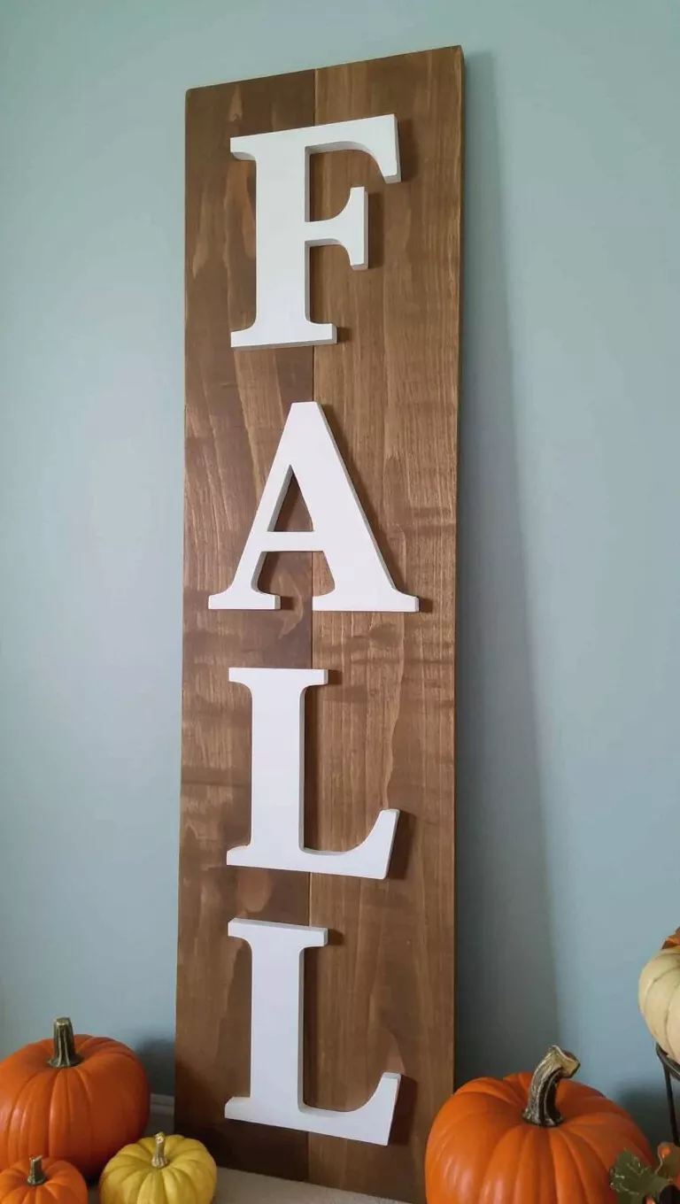 A photo of a tall vertical wooden sign with the word 'FALL' in large white letters leans against a light blue wall, with fall decor in the foreground.