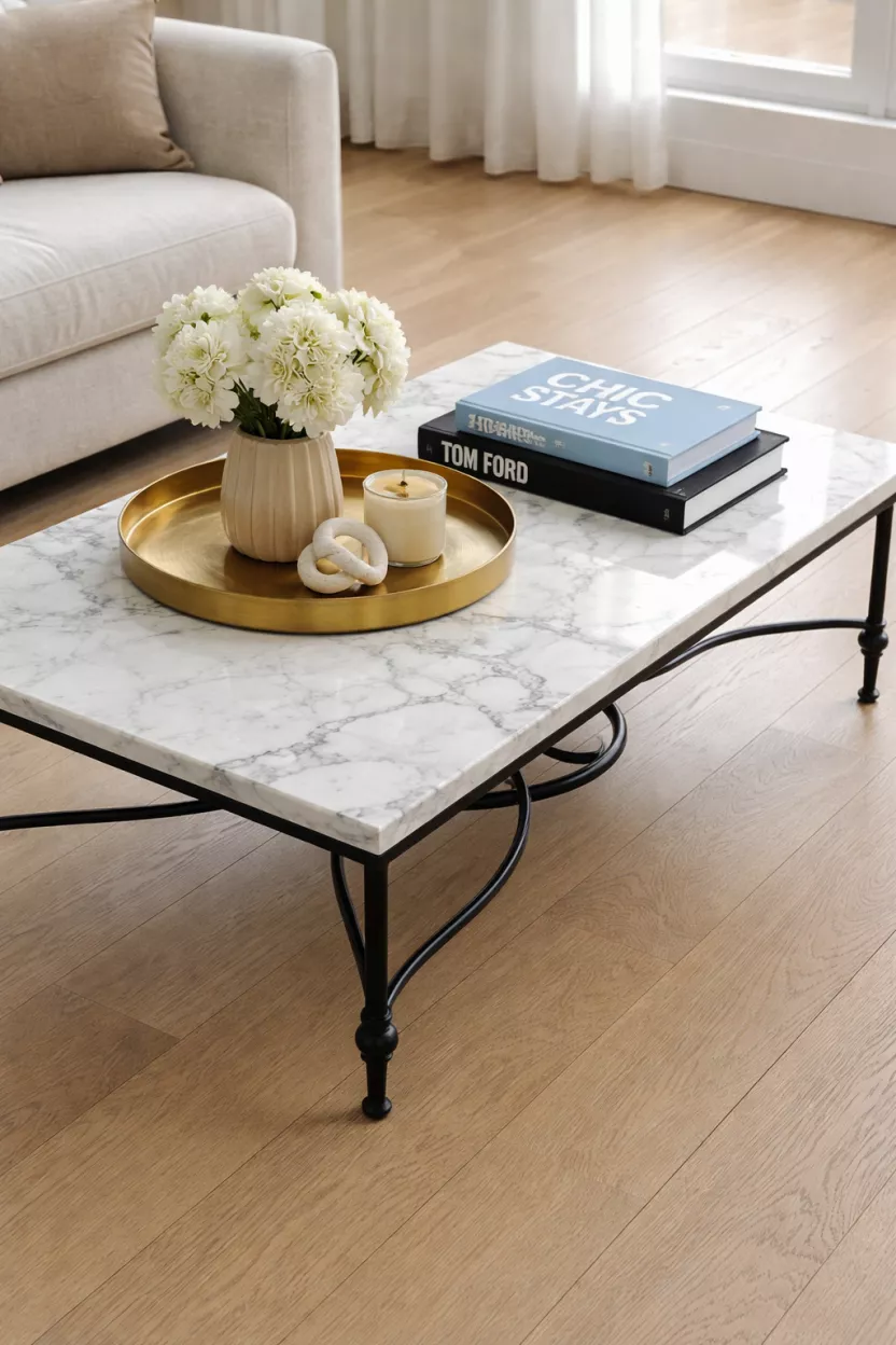 A realistic photo of a low white carrara marble coffee table with gray veining and a black wrought iron base, topped with a gold tray and thick art books, sitting on a light oak floor.