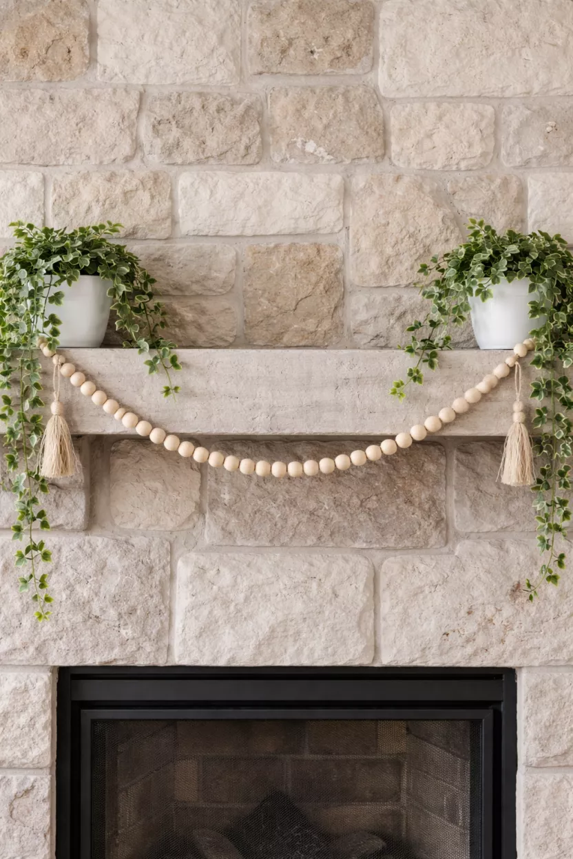 Beaded Wood Garland And Potted Ivy A realistic photo of a long light wood beaded garland draped across a stone mantel with two trailing green ivy plants in white pots on each end.