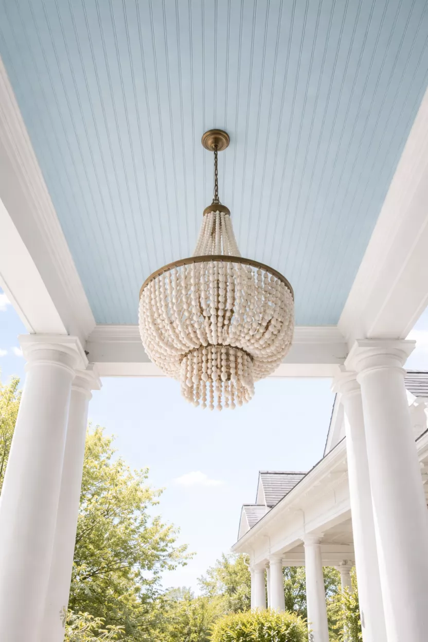 A realistic photo looking up at a porch ceiling painted a soft sky blue color, with a white beaded chandelier hanging from the center and white columns framing the view.