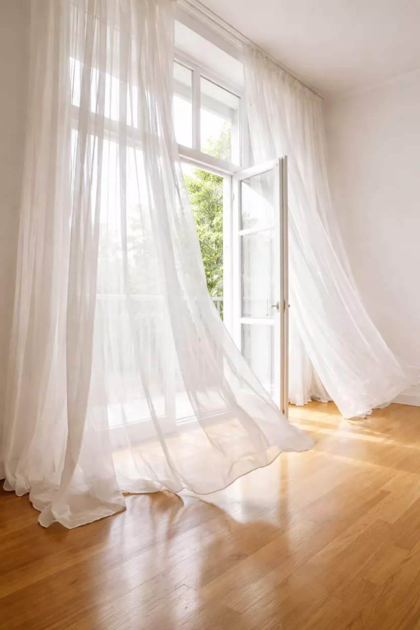 A realistic photo of long white sheer linen curtains blowing gently in a breeze by an open window, a polished oak wood floor reflects the natural light, a minimalist aesthetic with high ceilings and bright exposure.