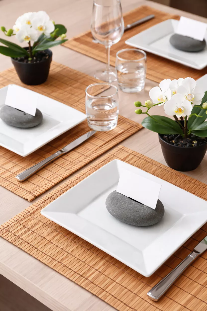 A realistic photo of bamboo placemats with smooth grey river stones used as place card holders, small white orchids in black pots, and clean white square plates.