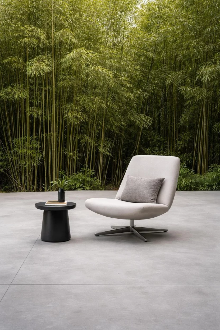A realistic photo of a massive, unadorned light gray concrete slab with a soft matte polish, featuring a single designer lounge chair and a small black side table against a backdrop of tall bamboo.