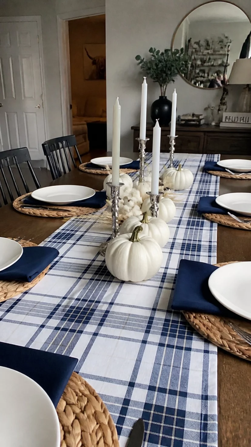 A super-realistic photo of a wooden dining table set with a navy blue and white plaid fabric runner, white ceramic dinner plates sit on top of woven seagrass placemats with navy blue cloth napkins, a centerpiece of white mini pumpkins and silver candlesticks runs down the middle, everyday social media post aesthetic, photorealistic, high-quality mobile photography, shot on an iPhone 14