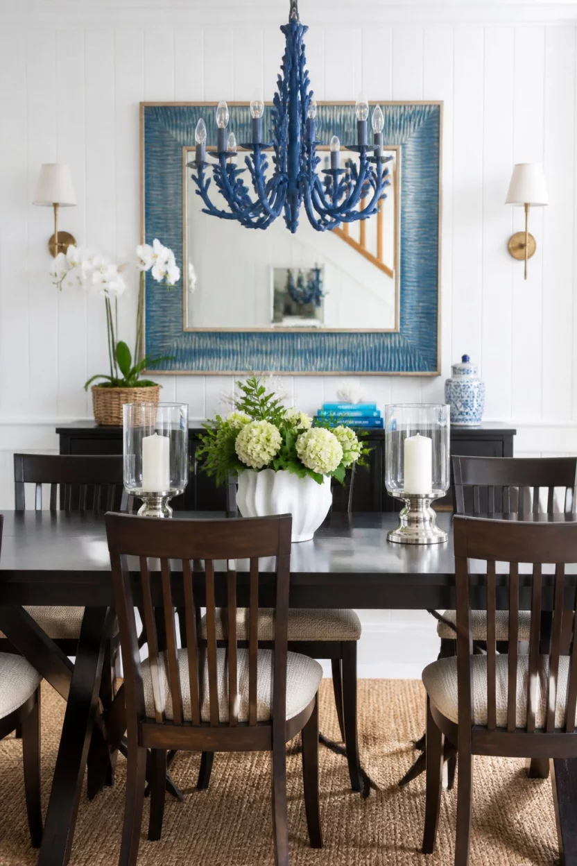 A realistic photo of a brightly lit dining room with white beadboard walls, a dark wood dining set, and vibrant blue accents in the mirror and chandelier.