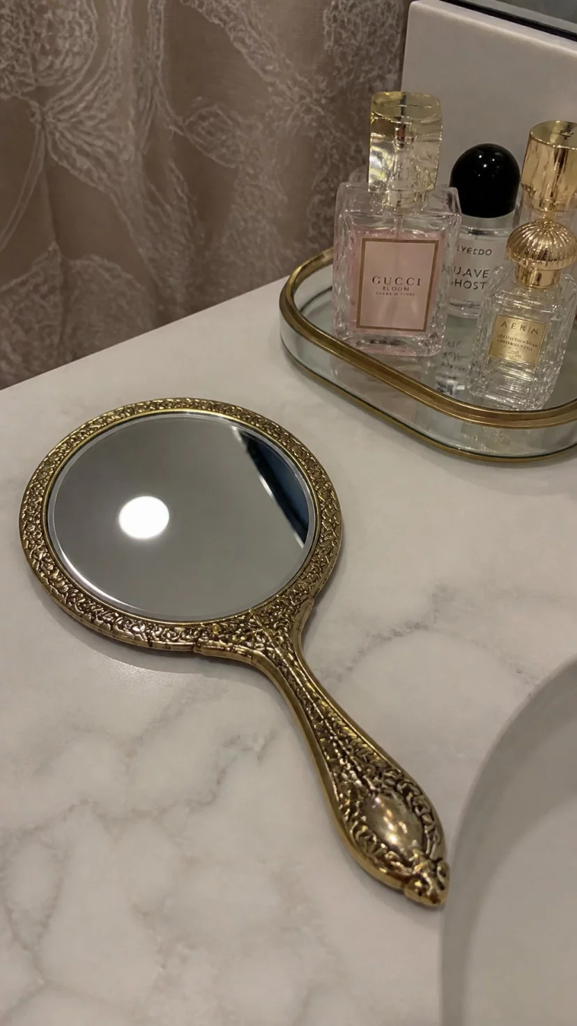 A super-realistic photo of a decorative antique brass hand mirror resting on a marble bathroom counter, the ornate handle featuring delicate engravings, the glass reflecting the ceiling light, a small glass tray of perfumes sitting next to it, the scene looking classic and feminine, a slight blur on the background shower curtain, everyday social media post aesthetic, photorealistic, high-quality mobile photography, shot on an iPhone 14