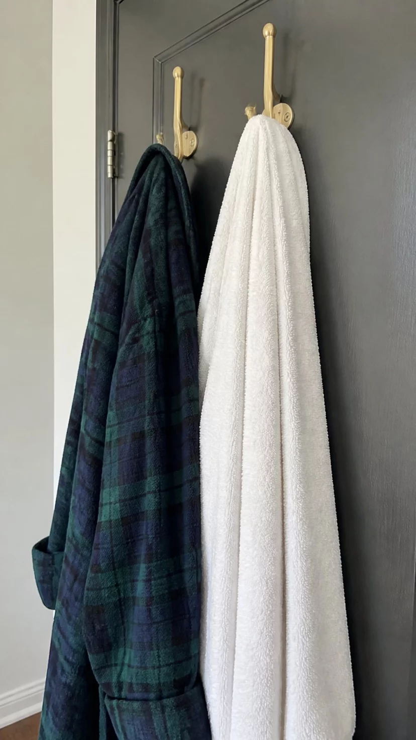 A super-realistic photo of two heavy brass hooks on the back of a bathroom door, a thick plaid flannel robe in navy and forest green hanging from one hook, the fabric looking soft and worn-in, the other hook holding a large white bath sheet, the door being a dark charcoal grey, soft light coming from the side, everyday social media post aesthetic, photorealistic, high-quality mobile photography, shot on an iPhone 14