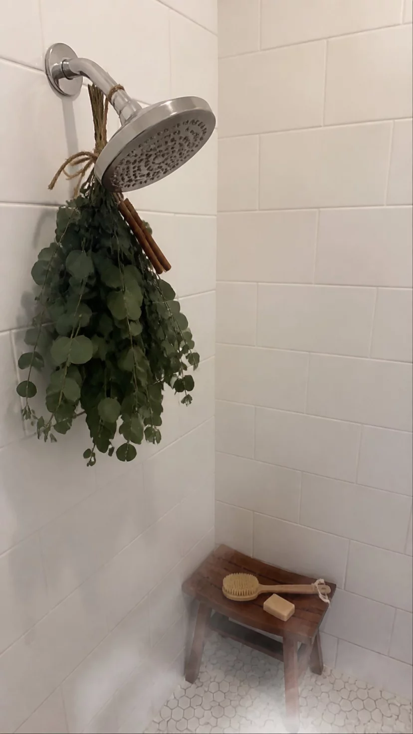 A super-realistic photo of a silver shower head with a lush bundle of dried green eucalyptus leaves and cinnamon sticks tied to the neck with rustic twine, steam gently rising from the shower floor below, the dark green leaves appearing slightly damp and vibrant against the white subway tiles, a small wooden stool visible in the corner of the walk-in shower, everyday social media post aesthetic, photorealistic, high-quality mobile photography, shot on an iPhone 14
