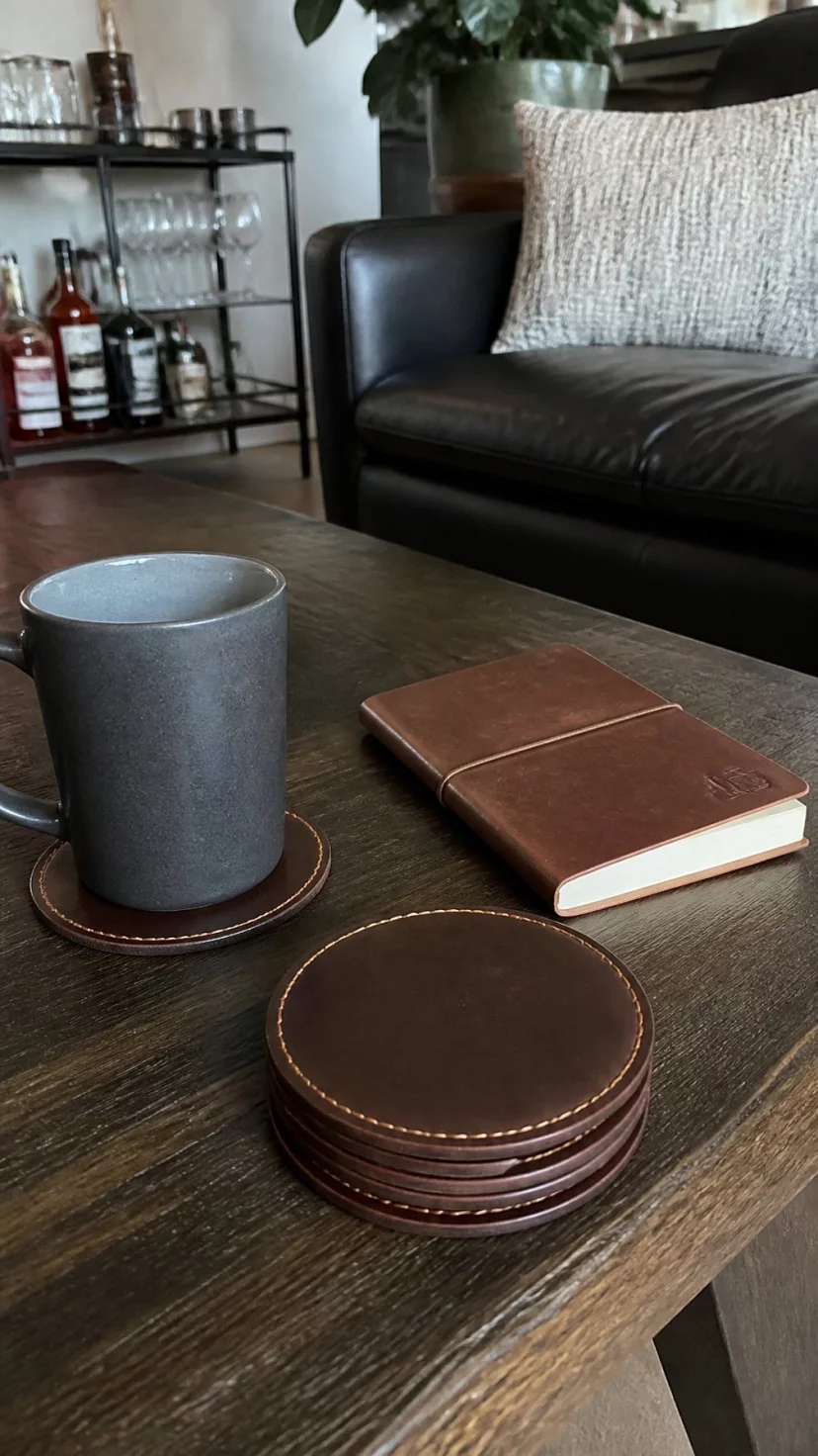 Leather Coasters And Heavy Mugs A super-realistic photo of a bachelor style living room coffee table with a set of dark brown leather coasters, a thick matte grey ceramic mug sits on one coaster, a small leather bound notebook is next to it, everyday social media post aesthetic, photorealistic, high-quality mobile photography, shot on an iPhone 14
