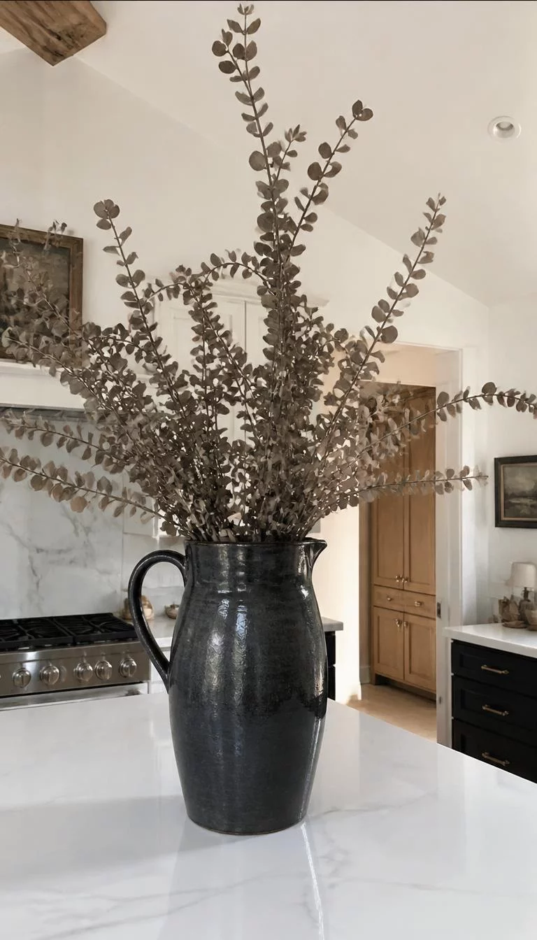A super-realistic photo of a kitchen island with a very large handmade ceramic pitcher in a charcoal glaze, filled with long branches of dried eucalyptus reaching toward the ceiling, everyday social media post aesthetic, photorealistic, high-quality mobile photography, shot on an iPhone 14