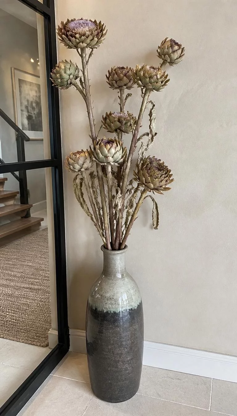 A super-realistic photo of a tall ceramic floor vase in a modern entryway, containing several tall dried artichoke stems with their purple tinged centers and rugged architectural leaves, leaning against a neutral plaster wall, everyday social media post aesthetic, photorealistic, high-quality mobile photography, shot on an iPhone 14