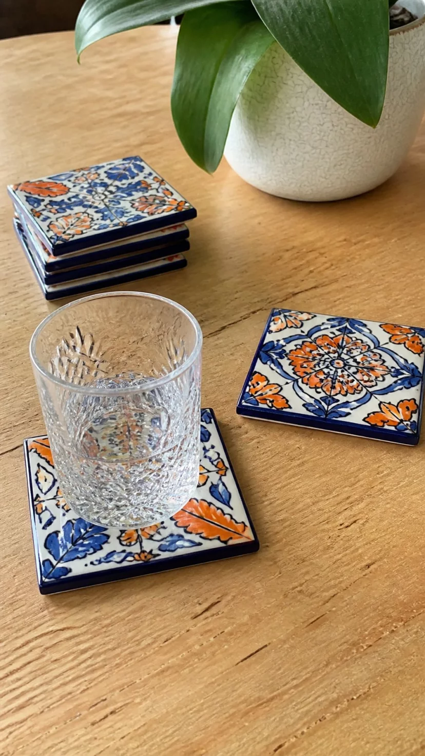 A super-realistic photo of a dining table surface, individual square ceramic tiles with hand painted navy and orange autumnal patterns used as drink coasters, a crystal glass sitting on one, artisanal and unique decor details, everyday social media post aesthetic, photorealistic, high-quality mobile photography, shot on an iPhone 14