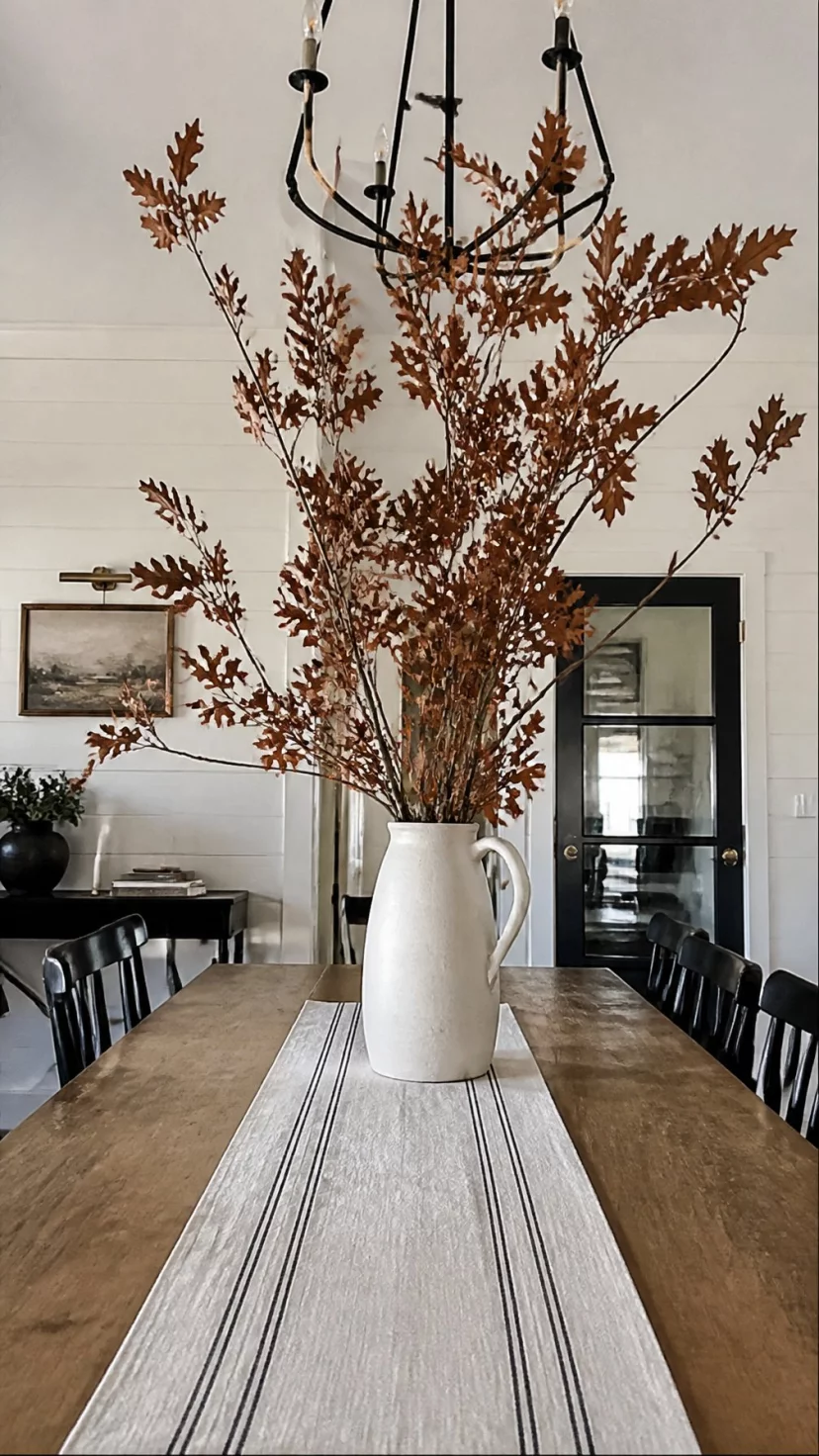 A super-realistic photo of a modern farmhouse dining room, a tall white ceramic pitcher in the center of the table, huge real oak branches with brown leaves reaching toward the ceiling, a dramatic and tall centerpiece, everyday social media post aesthetic, photorealistic, high-quality mobile photography, shot on an iPhone 14