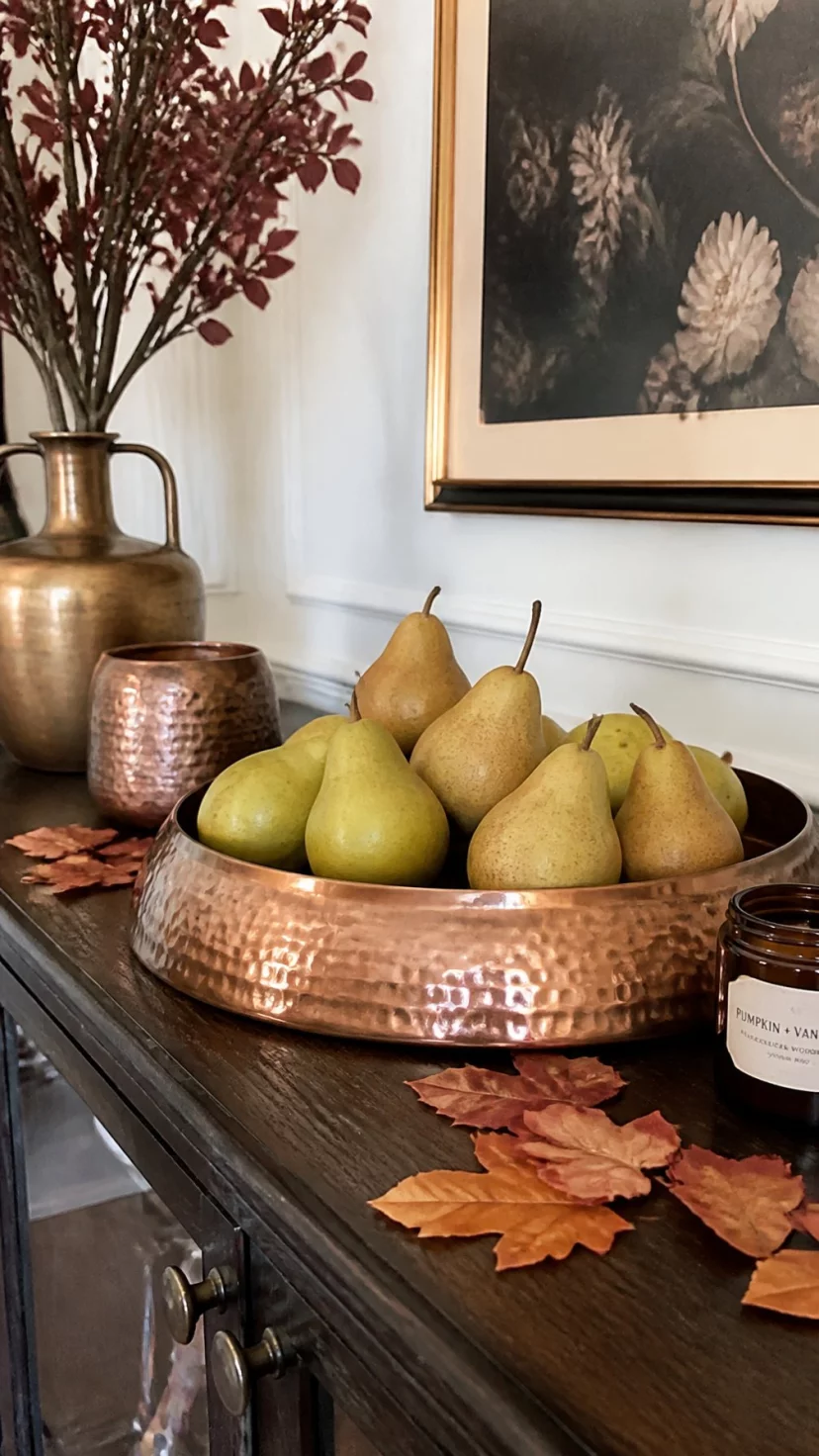 A super-realistic photo of a dining room sideboard, a large round beaten copper tray filled with ripe green and brown bosc pears, a few stray autumn leaves scattered around the base, warm metallic reflections, a still life inspired decor arrangement, everyday social media post aesthetic, photorealistic, high-quality mobile photography, shot on an iPhone 14