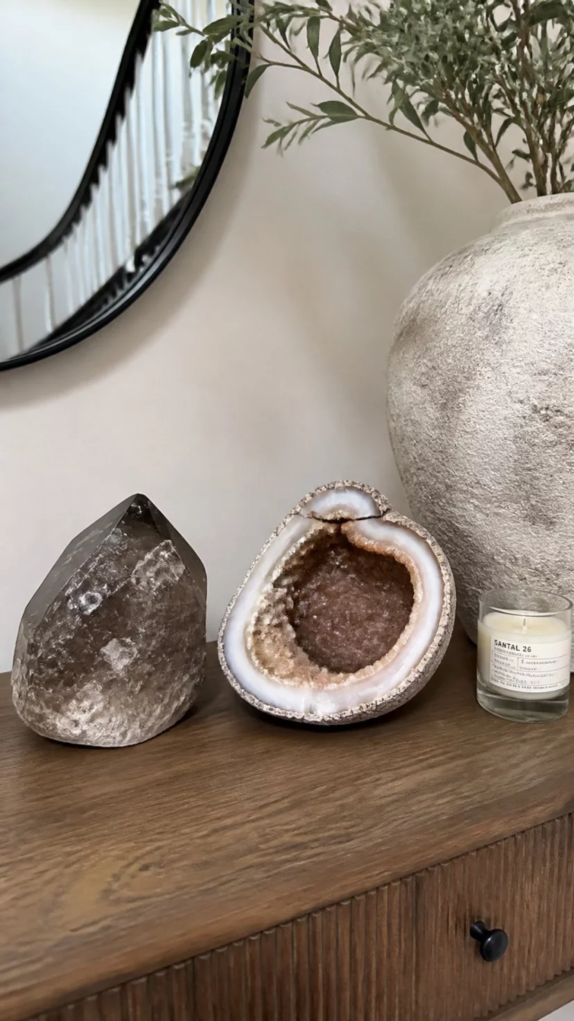 Smoky Quartz Crystals And Geodes A super-realistic photo of an entryway table featuring a large piece of smoky quartz and a cracked agate geode with brown and white interior details, the stones sit near a small glass candle jar, everyday social media post aesthetic, photorealistic, high-quality mobile photography, shot on an iPhone 14