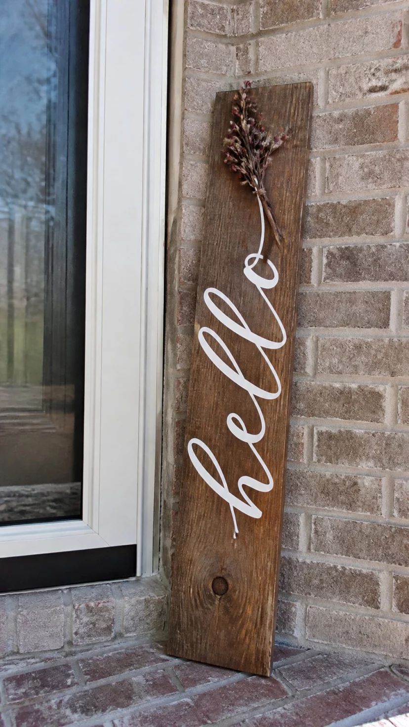 A super-realistic photo of a vertical raw wood plank leaning against a brick wall near a door, featuring a hand painted greeting in elegant white calligraphy with a small sprig of dried berries at the top, everyday social media post aesthetic, photorealistic, high-quality mobile photography, shot on an iPhone 14