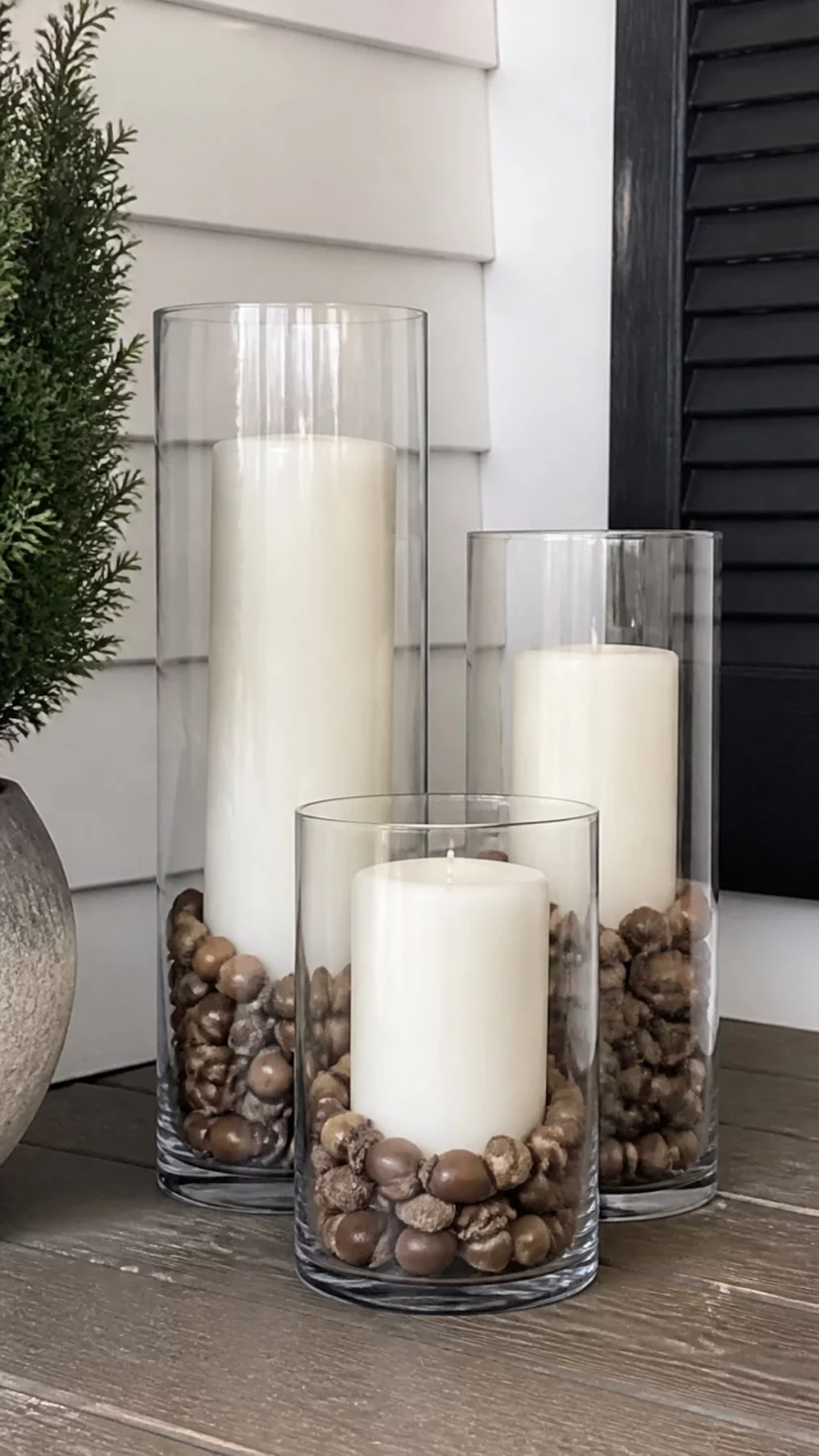A super-realistic photo of a set of three different sized clear glass hurricane vases on a porch, each containing a layer of acorns at the bottom and a tall white pillar candle, everyday social media post aesthetic, photorealistic, high-quality mobile photography, shot on an iPhone 14