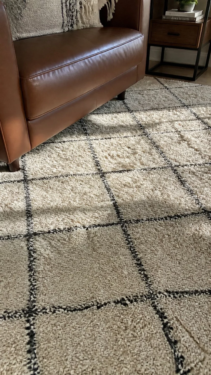 A super-realistic photo of a living room floor featuring a cream wool rug with thin charcoal grey lines in a diamond pattern, the edges of a cognac leather sofa touching the rug, sunlight hitting the wool fibers, everyday social media post aesthetic, photorealistic, high-quality mobile photography, shot on an iPhone 14