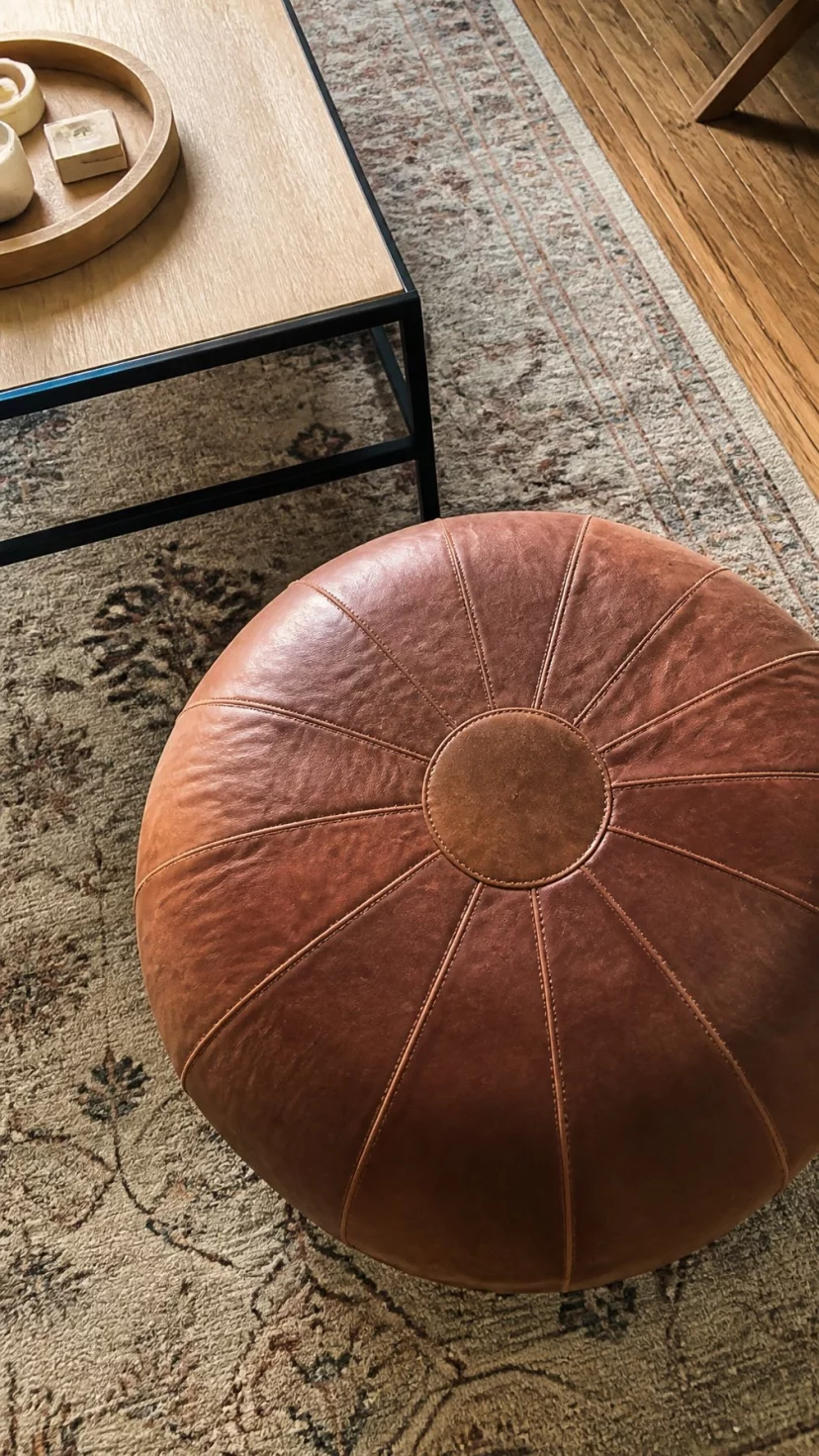 A super-realistic photo of a living room floor with a round cognac leather pouf, the leather showing natural wear and stitching details, a soft rug underneath, the edge of a coffee table nearby, everyday social media post aesthetic, photorealistic, high-quality mobile photography, shot on an iPhone 14