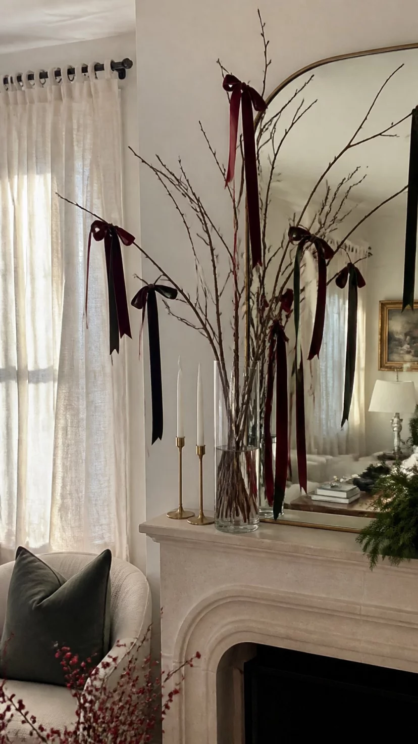 A super-realistic photo of a feminine living room showing a mantle with a tall glass vase filled with bare branches, several long velvet ribbons in deep burgundy and forest green tied in bows to the twigs, elegant and soft, hazy afternoon light, everyday social media post aesthetic, photorealistic, high-quality mobile photography, shot on an iPhone 14