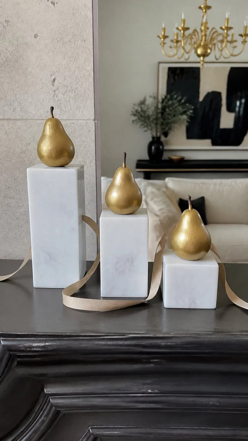 A super-realistic photo of a luxury living room where a dark mantle holds three white marble pedestals of varying heights, each pedestal supports a single gold-painted pear, a thin silk ribbon weaves between them, high-end and sophisticated, soft studio-style lighting, everyday social media post aesthetic, photorealistic, high-quality mobile photography, shot on an iPhone 14