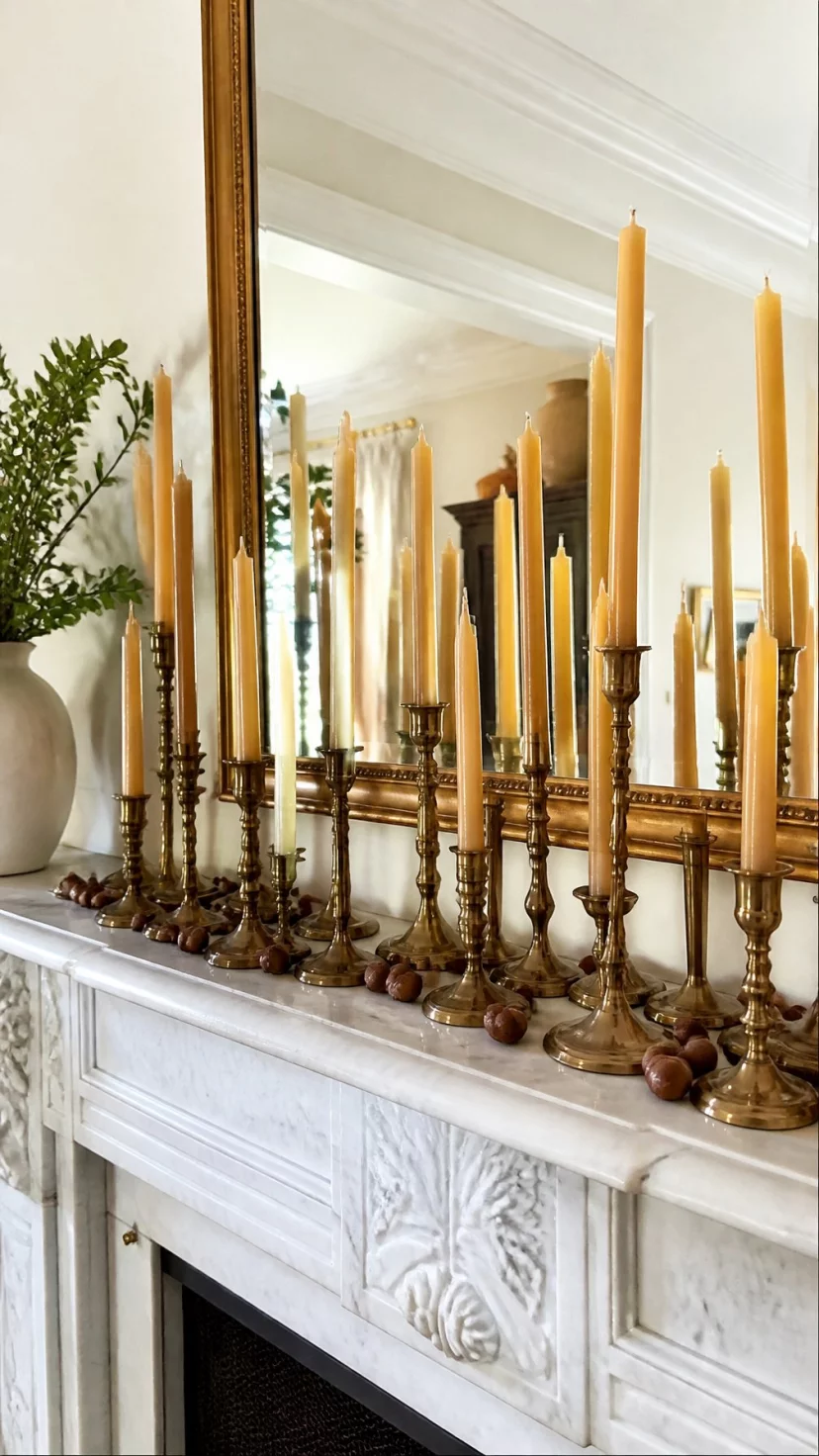 A super-realistic photo of a bright living room showing a white marble mantle decorated with a collection of twenty antique brass candlesticks of varying heights holding cream beeswax tapers, small acorns scattered at the base of the metal holders, a large gold-framed mirror reflecting the warm room, elegant and cozy atmosphere, everyday social media post aesthetic, photorealistic, high-quality mobile photography, shot on an iPhone 14