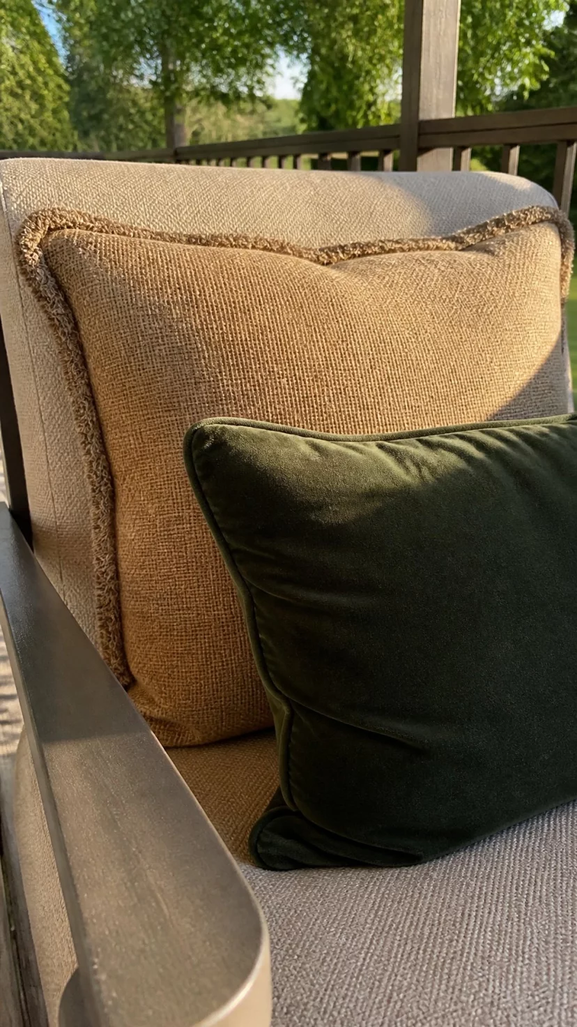 A super-realistic photo of a close up on an outdoor armchair with two pillows, one is made of rough tan burlap and the other is a dark green velvet, the textures provide a contrast, a wooden porch railing is visible in the background, warm afternoon light, everyday social media post aesthetic, photorealistic, high-quality mobile photography, shot on an iPhone 14