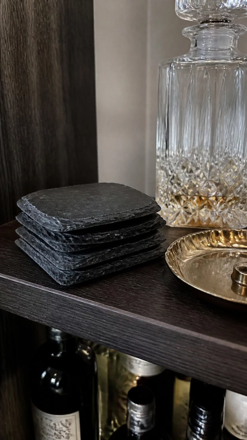 A super-realistic photo of a bar shelf featuring a stack of rough-edged dark gray slate coasters next to a crystal decanter and a small brass tray, everyday social media post aesthetic, photorealistic, high-quality mobile photography, shot on an iPhone 14