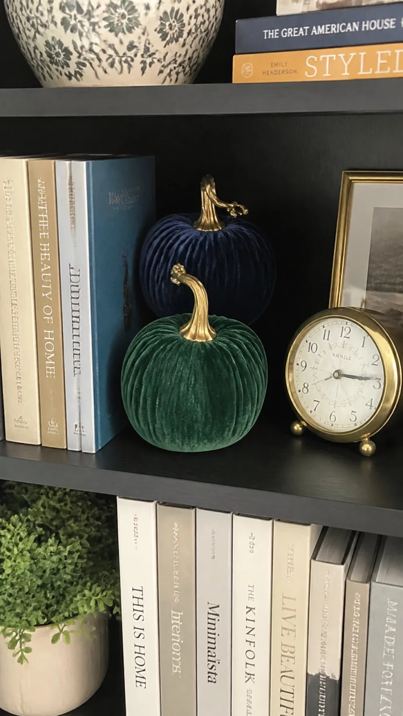 A super-realistic photo of a bedroom bookshelf featuring soft velvet pumpkins in emerald green and navy blue nestled between rows of hardback books and a small gold clock, everyday social media post aesthetic, photorealistic, high-quality mobile photography, shot on an iPhone 14
