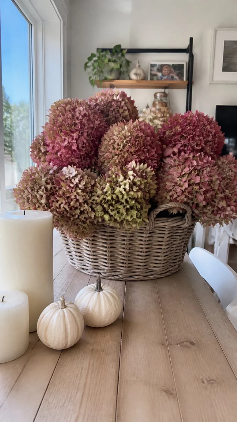 Dried Hydrangeas In Woven Baskets A super-realistic photo of a light-colored wooden table with a large round woven wicker basket holding a massive bunch of dried hydrangea blooms in shades of dusty rose and faded green, a few cream-colored candles and small white gourds nearby, soft natural light from a large window, everyday social media post aesthetic, photorealistic, high-quality mobile photography, shot on an iPhone 14