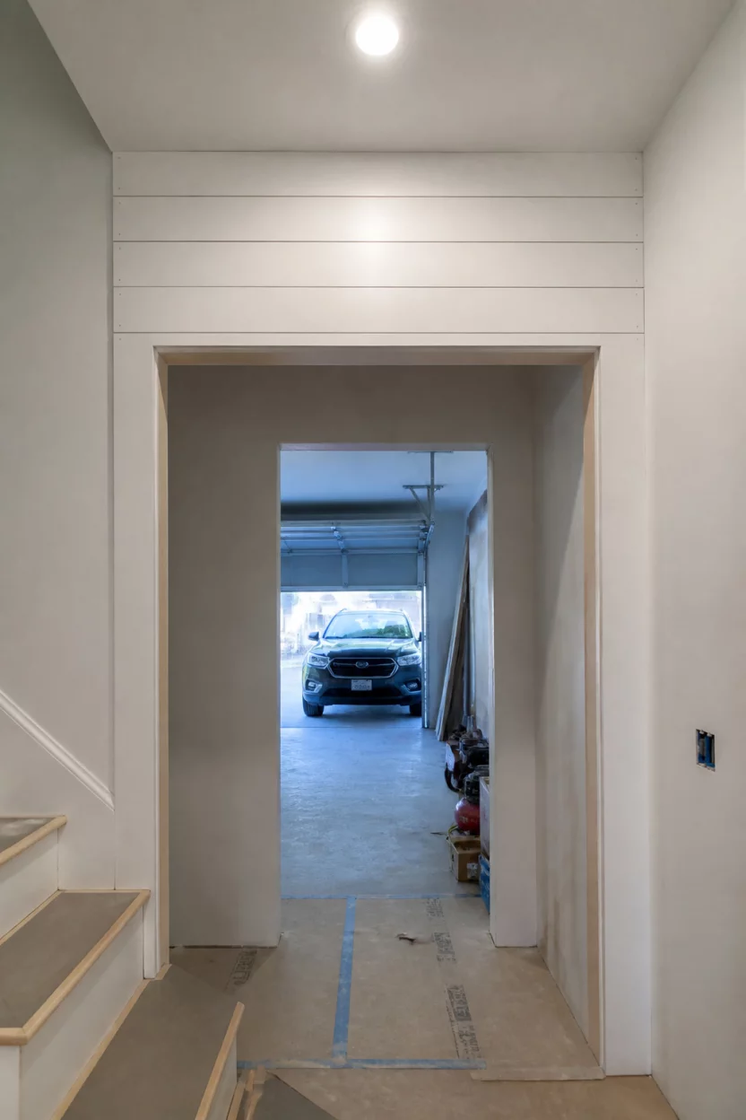 Shiplap Construction Zone A realistic photo of a brightly lit hallway entry under construction featuring white shiplap installation above a door opening that leads to a garage with a car visible in the distance.