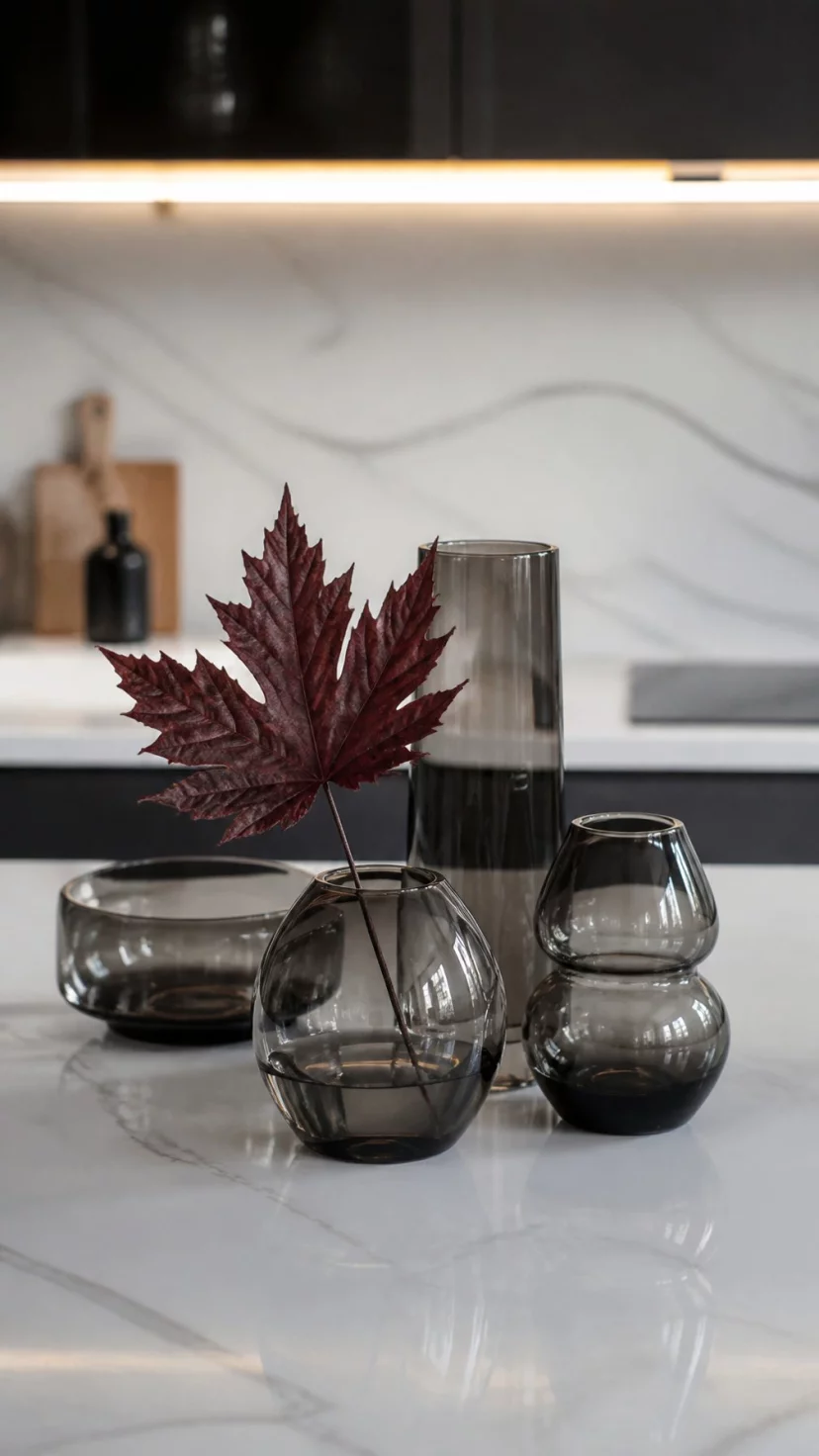 A super-realistic photo of a cluster of smoked grey glass vases on a white marble kitchen island, one vase contains a single dark red maple leaf, the glass is semi-transparent and moody, everyday social media post aesthetic, photorealistic, high-quality mobile photography, shot on an iPhone 14