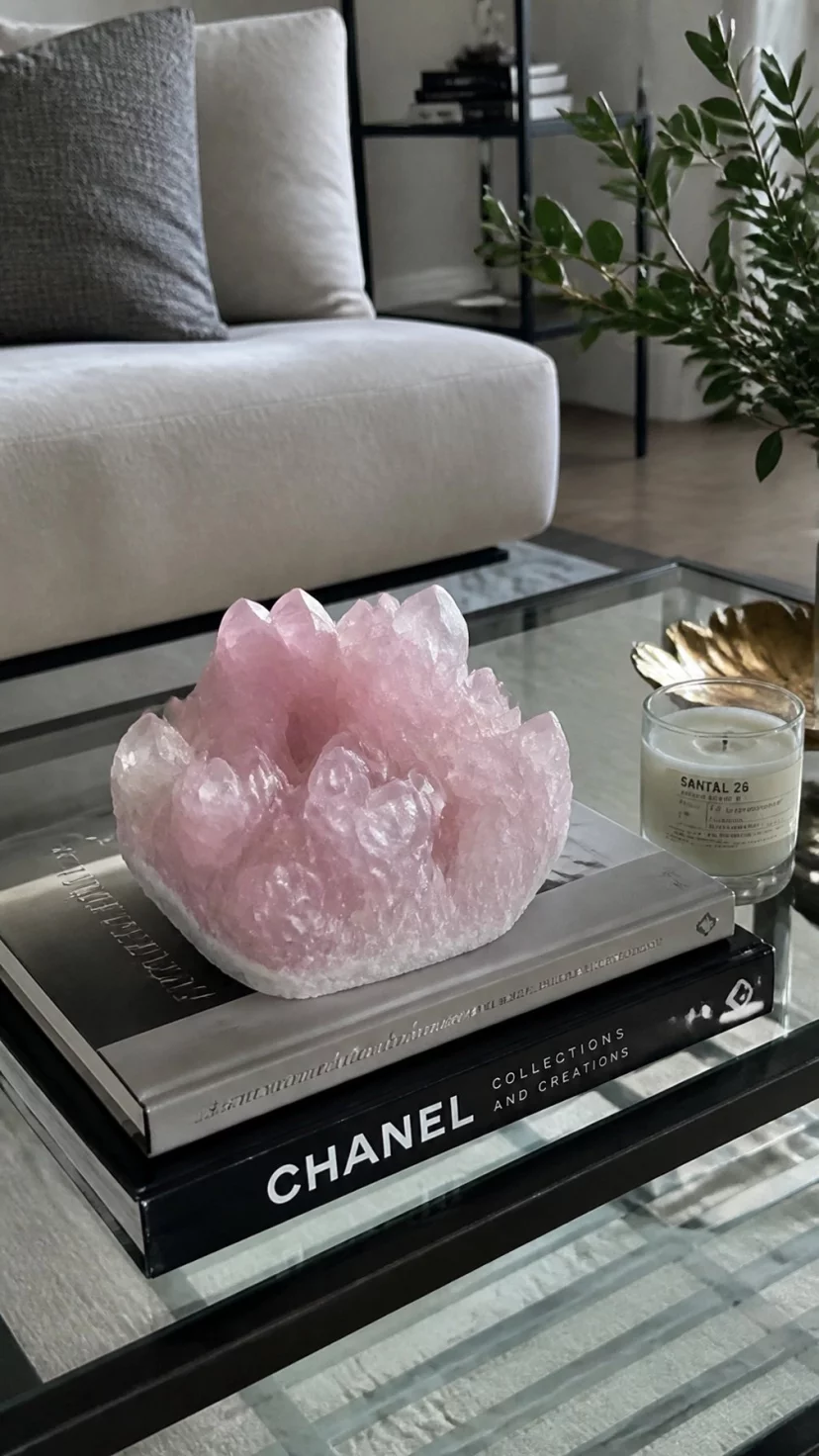 A super-realistic photo of a coffee table featuring a large rose quartz crystal cluster resting on top of a stack of art books, a small scented candle nearby, the natural facets of the crystal reflecting soft indoor light, everyday social media post aesthetic, photorealistic, high-quality mobile photography, shot on an iPhone 14