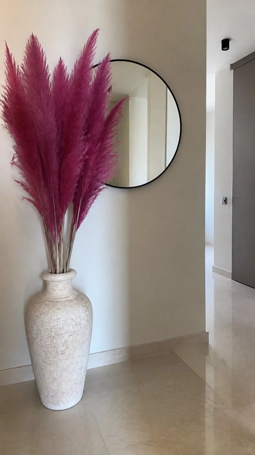 A super-realistic photo of a floor vase in a minimalist hallway containing tall stalks of fuchsia dried pampas grass, the feathery plumes reaching toward the ceiling, a simple circular mirror on the wall behind the arrangement, everyday social media post aesthetic, photorealistic, high-quality mobile photography, shot on an iPhone 14