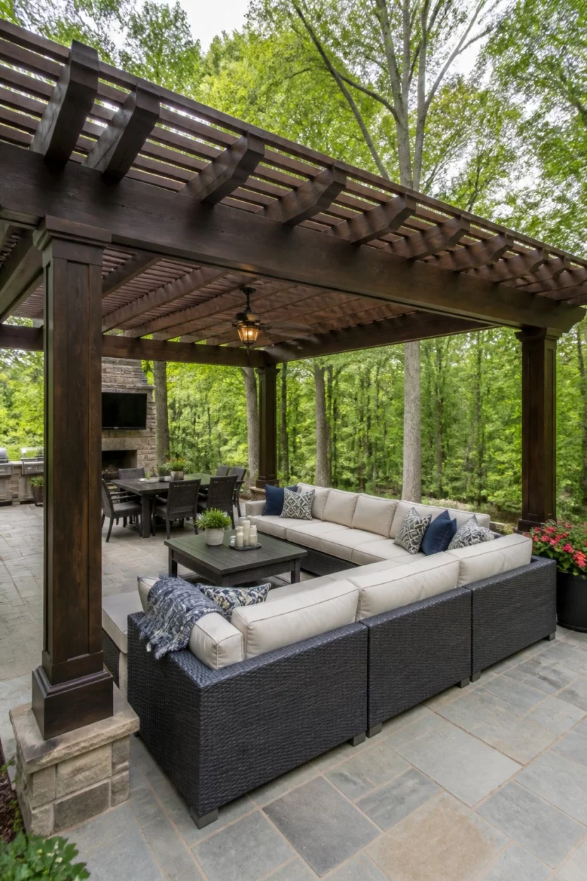 Grand U Shaped Sectional Shelter A realistic photo of a substantial dark wood pergola with thick posts covering a large stone patio that holds a massive U shaped wicker sectional sofa, surrounded by tall leafy trees in a private backyard.