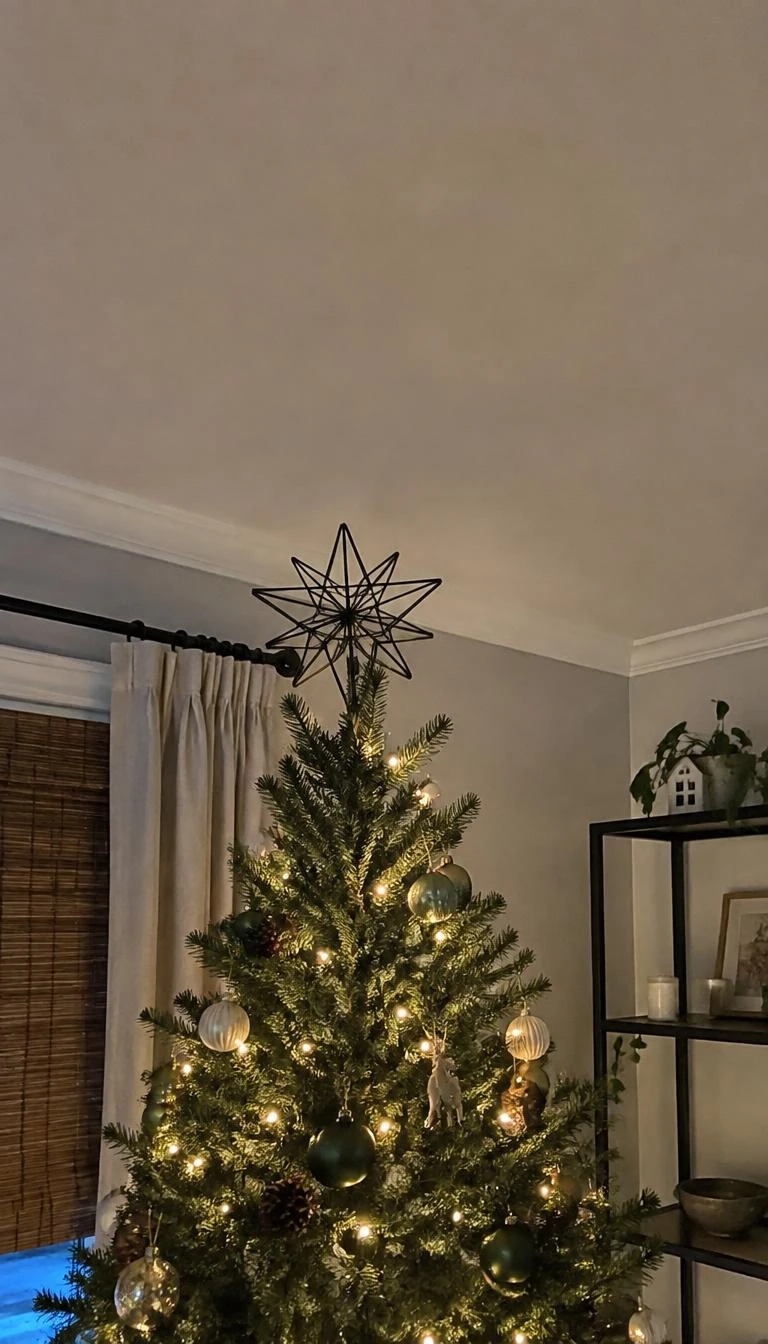 A candid, realistic iPhone photograph of a living room with a Christmas tree, showing an open black wire geometric star tree topper against a neutral ceiling, warm soft lighting throughout the room.