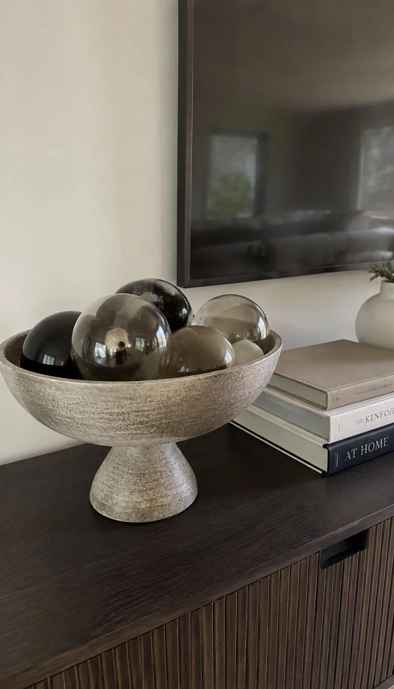 A candid, realistic iPhone photograph of a living room sideboard featuring a footed matte stoneware compote bowl filled with smoked black glass and semi transparent baubles, sitting beside a small stack of cloth bound books.