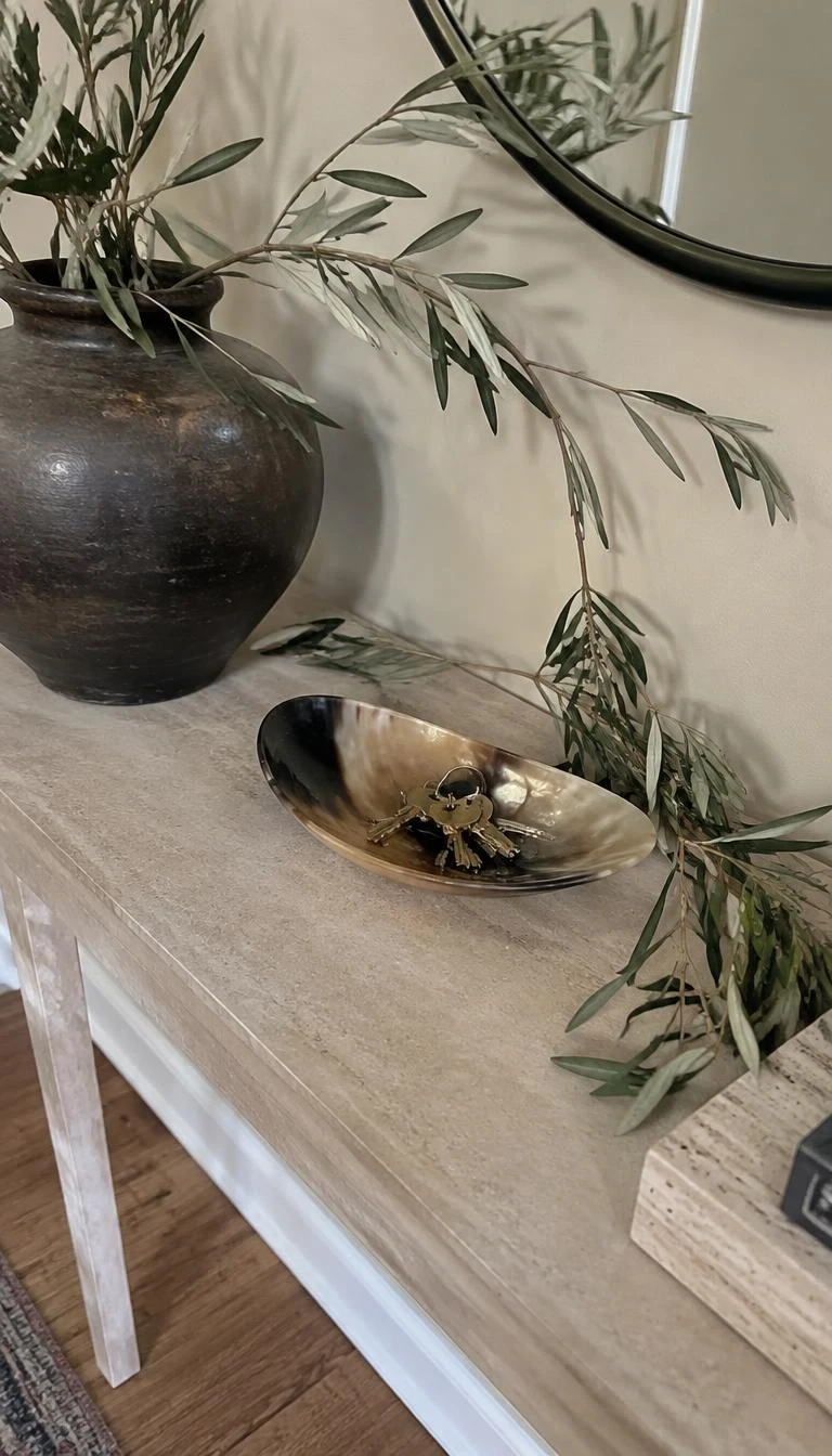 Weave Wild Olive Branches Through Horn Catchall Bowls A candid, realistic iPhone photograph of an entryway table, showing wild silver-green olive branches woven around a polished natural horn catchall dish containing brass keys, positioned on a light stone console table.