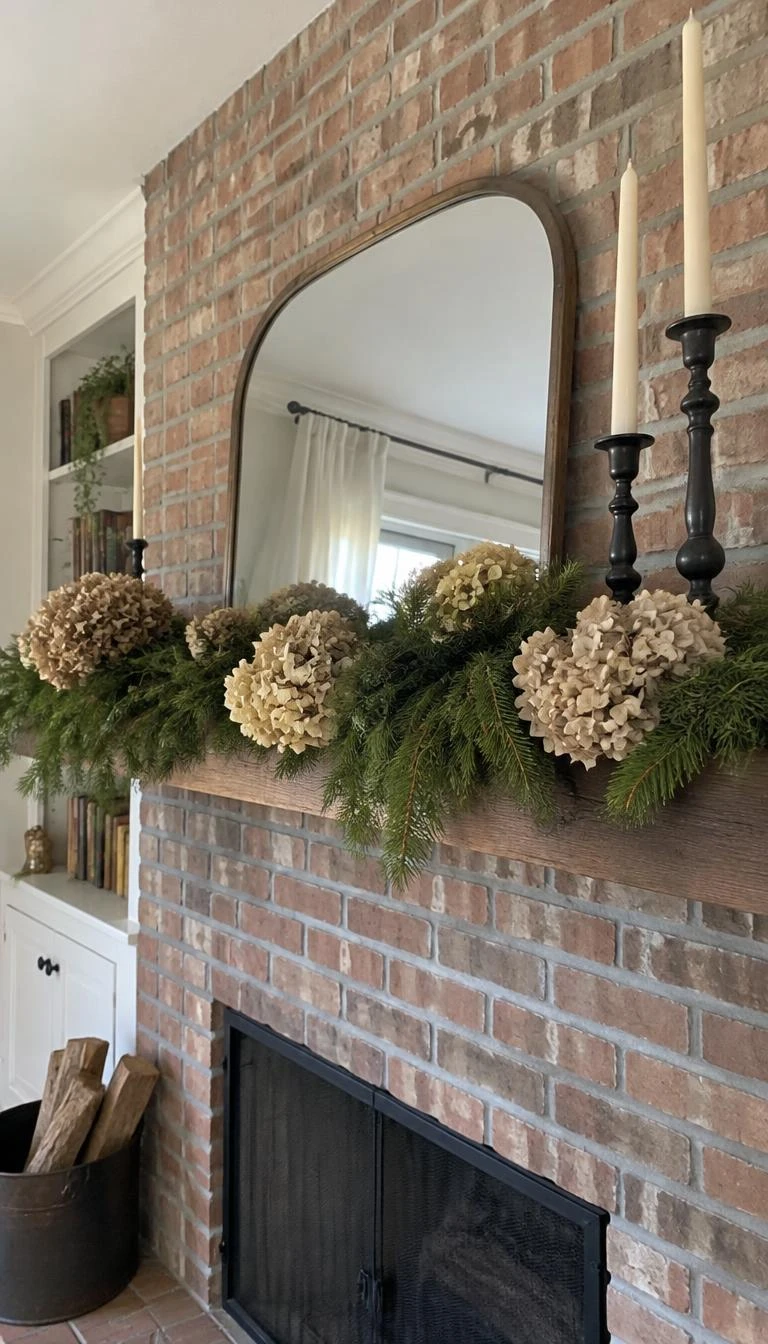 A candid, realistic iPhone photograph of a brick fireplace mantel, decorated with pale tan dried hydrangea heads tucked into freshly cut eastern white pine branches, cast iron candleholders nearby, soft natural daylight, simple home styling.