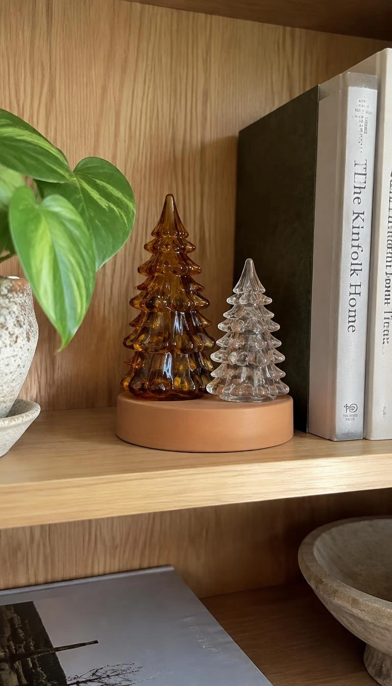 Rest Handblown Glass Trees On Small Pedestals A candid, realistic iPhone photograph of a light oak bookcase, showing two handblown amber and clear glass evergreen trees resting on an upturned terracotta saucer, gentle daylight, clear reflections