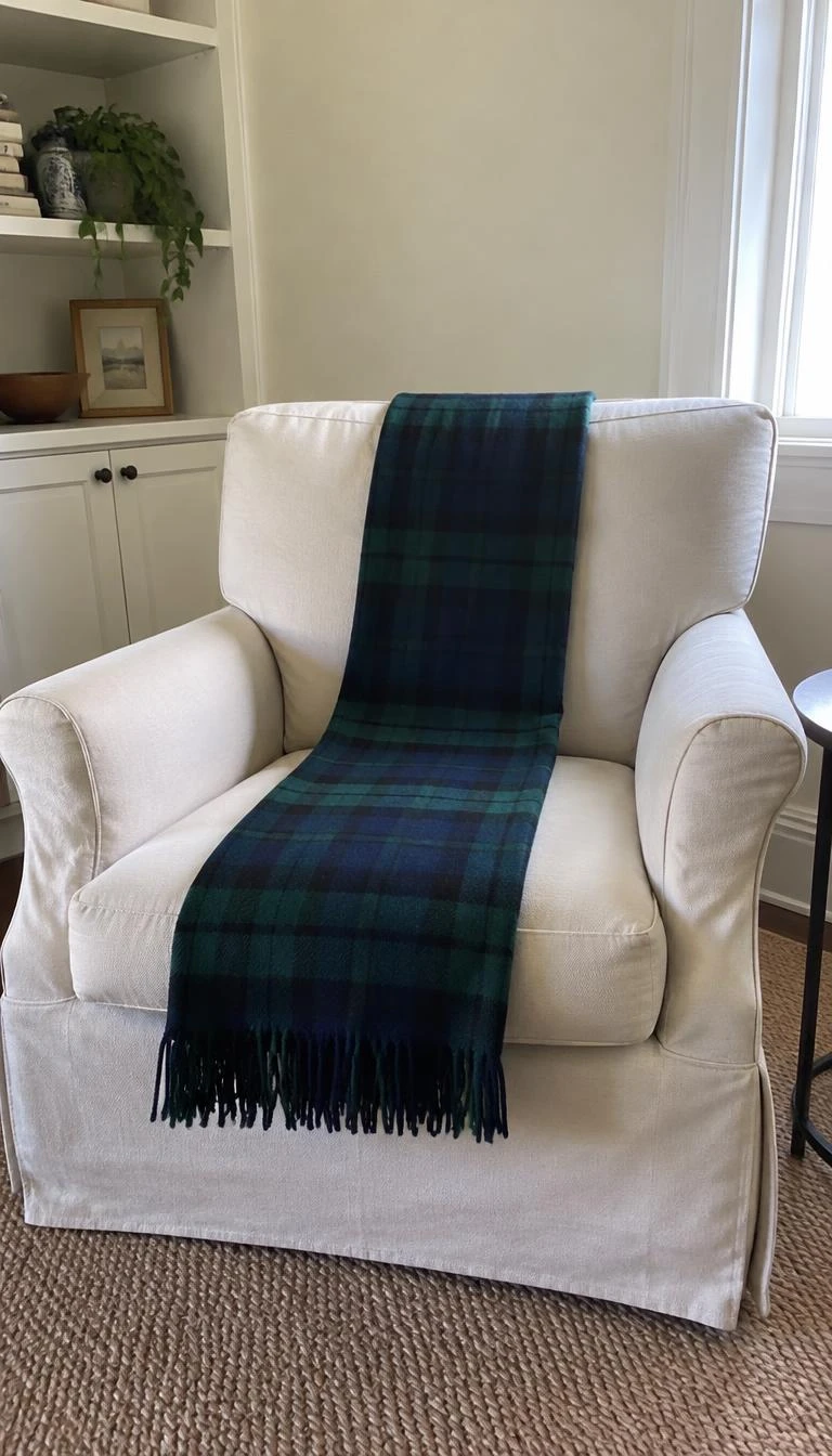 A candid, realistic iPhone photograph of a living room armchair, featuring a cream linen slipcovered chair draped with a green and navy tartan throw folded lengthwise down the center cushion.