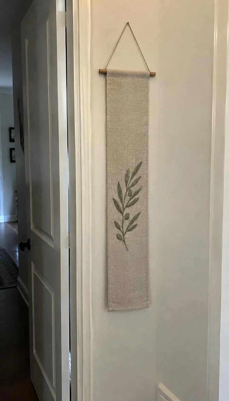 A candid, realistic iPhone photograph of an interior hallway door frame, showing a narrow oatmeal linen banner embroidered with a subtle olive leaf sprig in matte green cotton thread hanging from a slender wooden dowel.