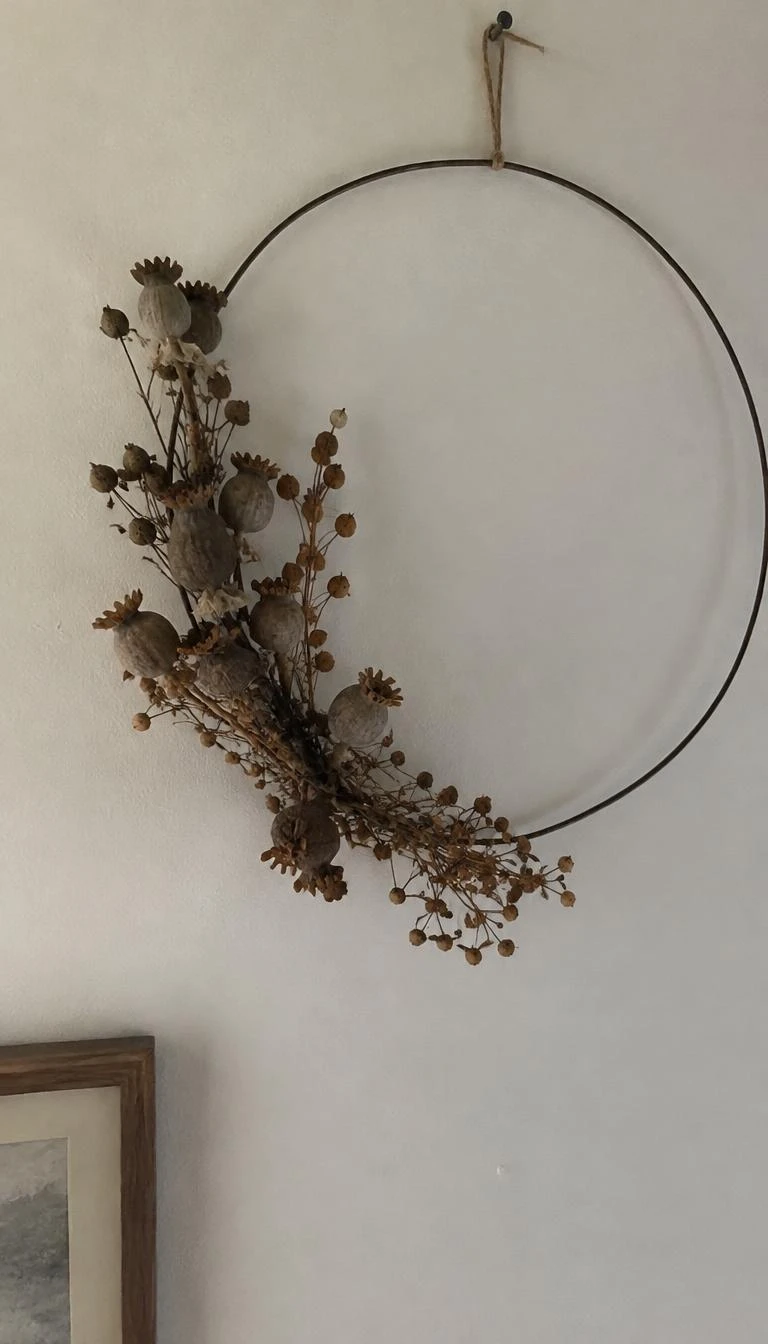 Display Dried Seed Pods on Flat Iron Wreaths A candid, realistic iPhone photograph of an interior white wall, a slender dark iron hoop wreath decorated asymmetrically with dried poppy heads and brown seed pods, soft natural light
