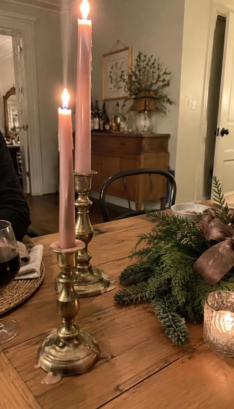 A candid, realistic iPhone photograph of a dining room table setup, aged heavy brass candlesticks holding slender pale pink beeswax taper candles, wax drips on a natural wood surface, soft ambient holiday lighting, casual angle