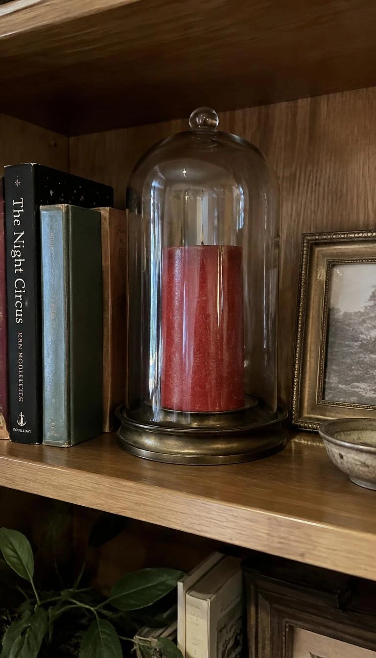 Fill Glass Cloches With Red Wax Candles And Brass Trimmings A candid, realistic iPhone photograph of a wooden bookshelf styling featuring a tall clear glass cloche over a thick red wax pillar candle resting in an old brass saucer base