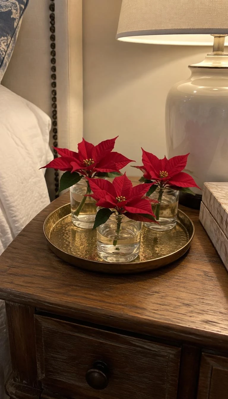 Use A Hammered Brass Tray For Poinsettia Cuttings A candid, realistic iPhone photograph of a bedroom nightstand showing a small round hammered brass tray holding three short stem red poinsettia blossoms in water inside clear glass votives